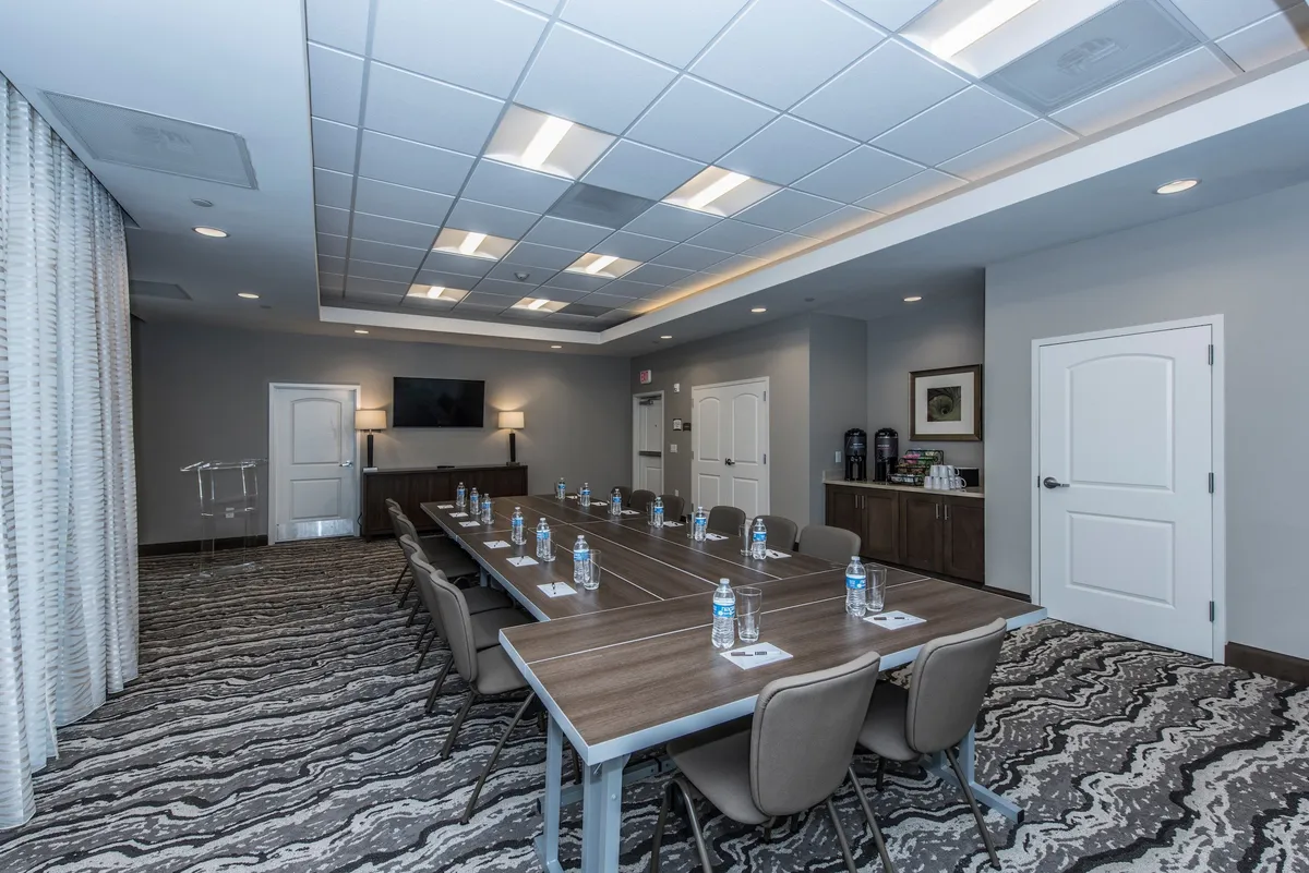 A contemporary business center with a long conference table set up for meetings, water bottles, a television screen, and a coffee station. The space is ideal for business meetings, workshops, and productive work sessions away from home.