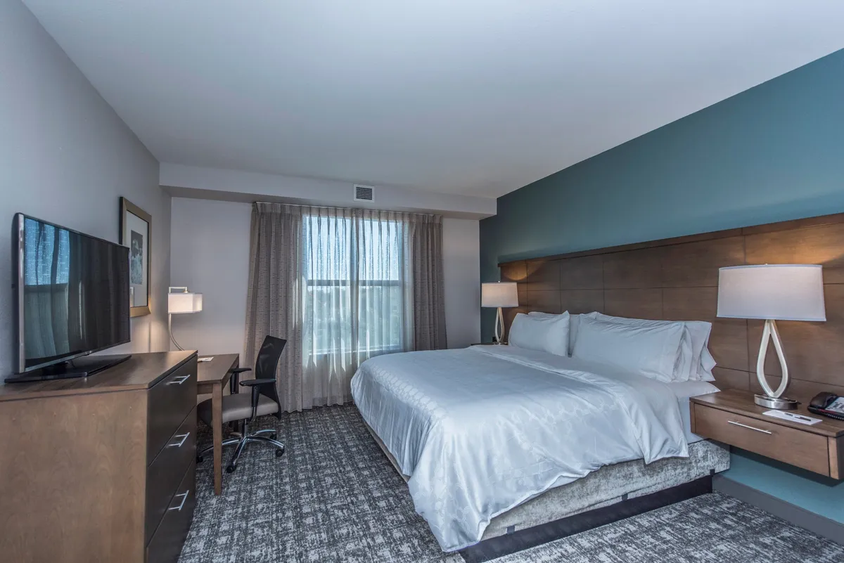 A bright and restful bedroom featuring a neatly made queen bed, a desk for personal work, a television for entertainment, and a sunlit window that enhances the feeling of spaciousness. The decor is understated and modern, emphasizing comfort and relaxation.