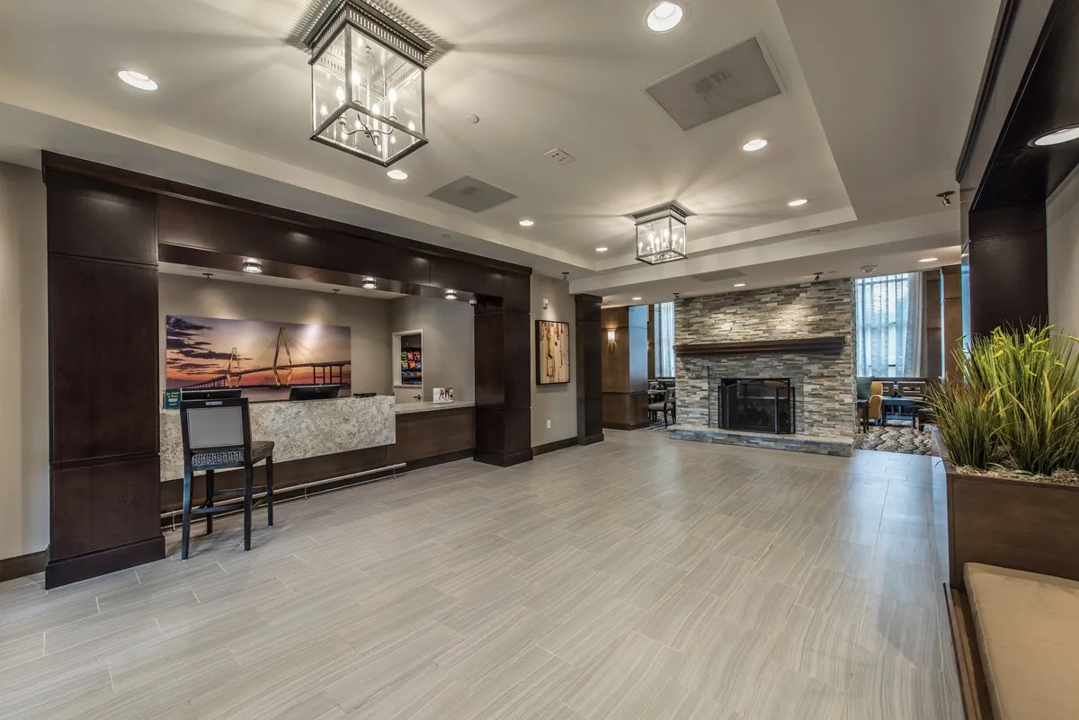 This image features the welcoming reception desk, a cozy fireplace, stylish artwork on the wall, and lush potted plants. Guests are shown checking in, socializing by the fireplace, and relaxing in comfortable lounge seating, creating a lively and inviting atmosphere.
