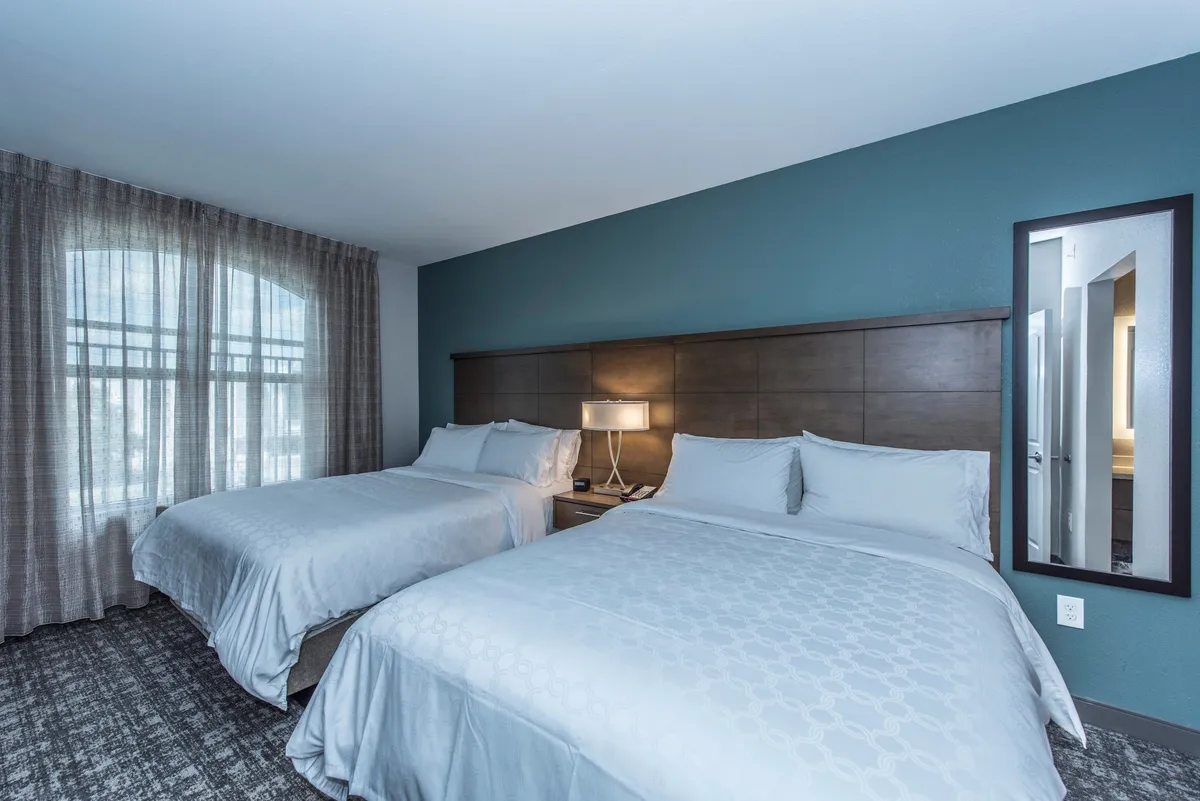 A cozy bedroom with two neatly arranged double beds separated by a nightstand with a lamp, a large window with flowing curtains, and a mirror. The room appears airy, well-lit, and inviting, suitable for families or multiple guests.