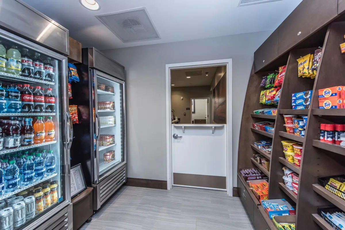 The image presents a well-stocked refrigerated drink section and a nearby snack aisle. Guests are seen grabbing refreshments, purchasing snacks, and casually interacting with staff, conveying a sense of convenience and hospitality.