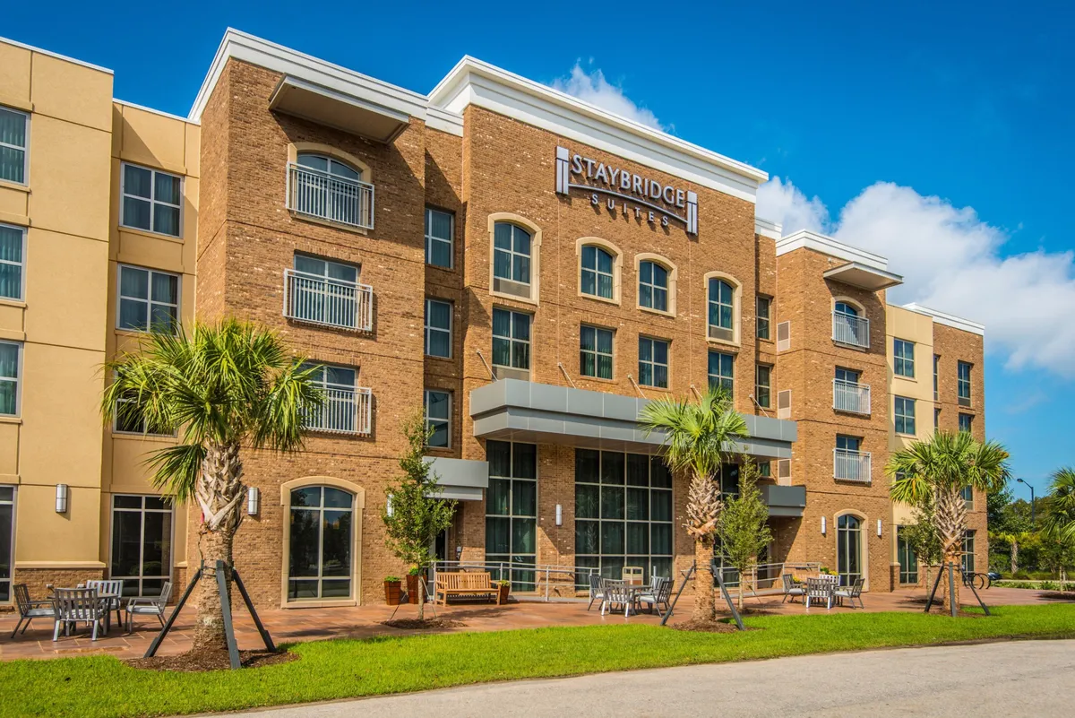 The image displays the Staybridge Suites sign, palm trees, and a comfortable outdoor seating area. The scene suggests easy access from the parking area to outdoor amenities, with guests relaxing and enjoying the sunshine.